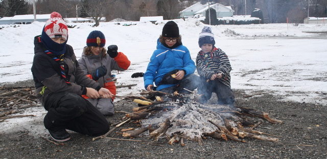 4 garçons entre 9 et 11 ans sont agenouillés autour d'un feu presqu'éteint, lors d'un camp, en hiver. De la neige est visible en arrière plan.